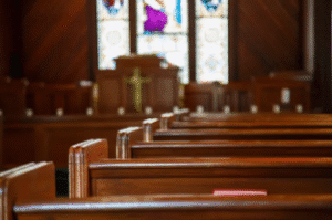 Empty wooden pews inside a church with a cross and stained glass windows.