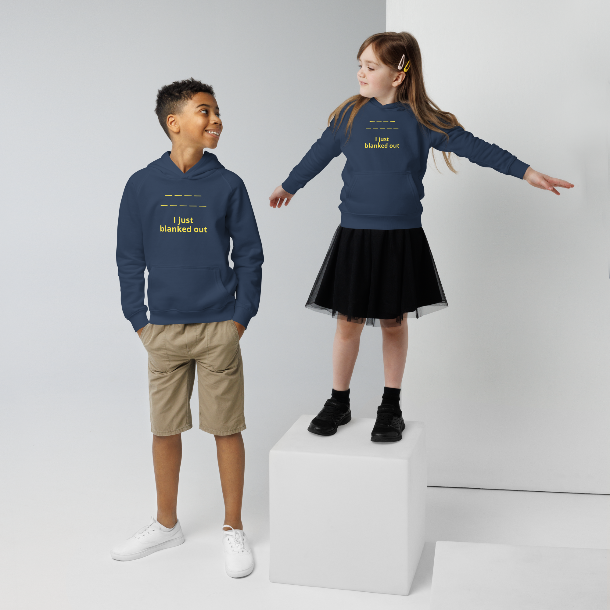 Two children wearing navy hoodies with yellow text, one standing on a white cube.
