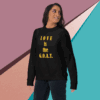 unisex-premium-sweatshirt-black-front-68f061abc22c8 Woman wearing a black sweatshirt with 'LOVE IS the G.O.A.T.' text.