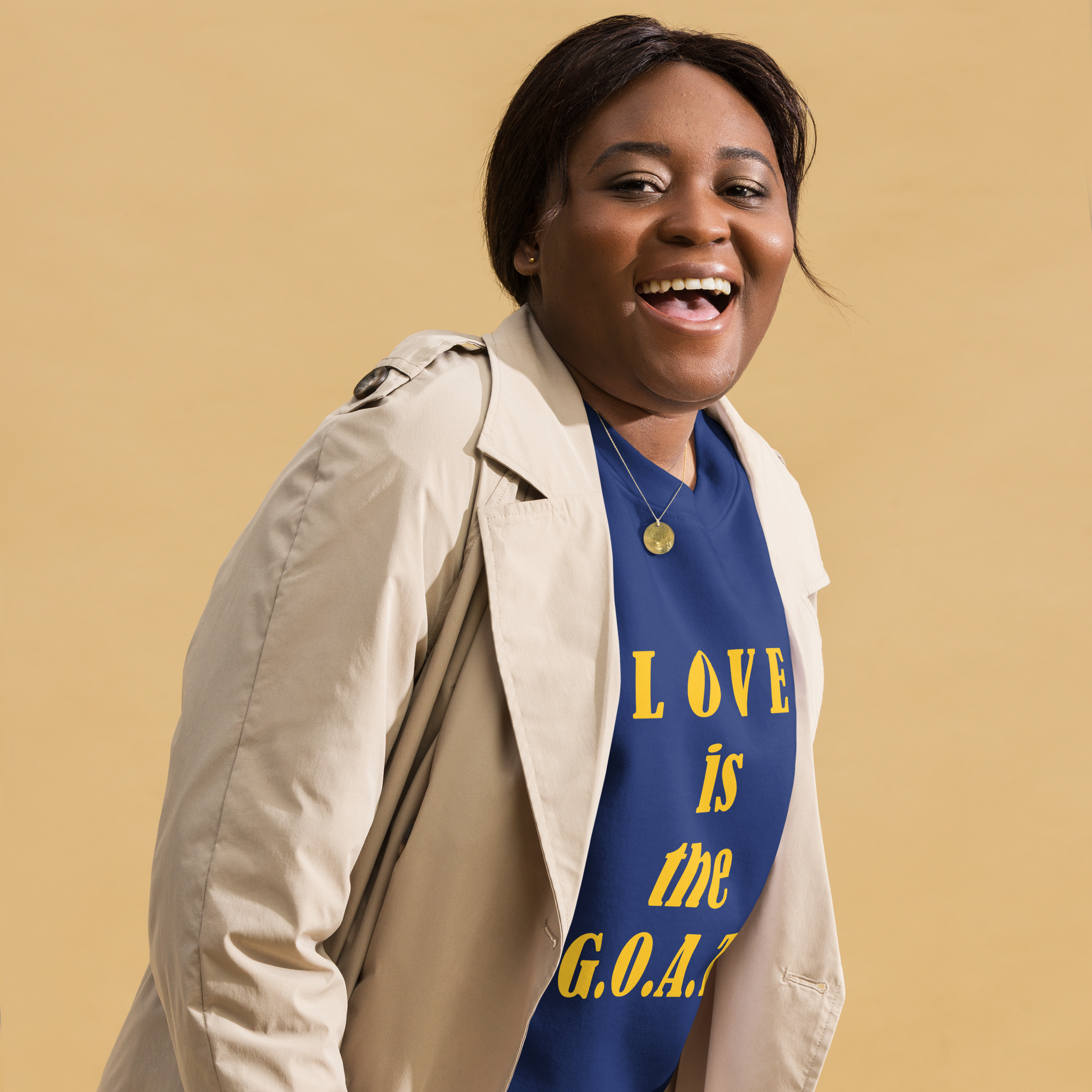 Smiling woman wearing a blue shirt with "LOVE is the G.O.A.T" text and a beige coat.