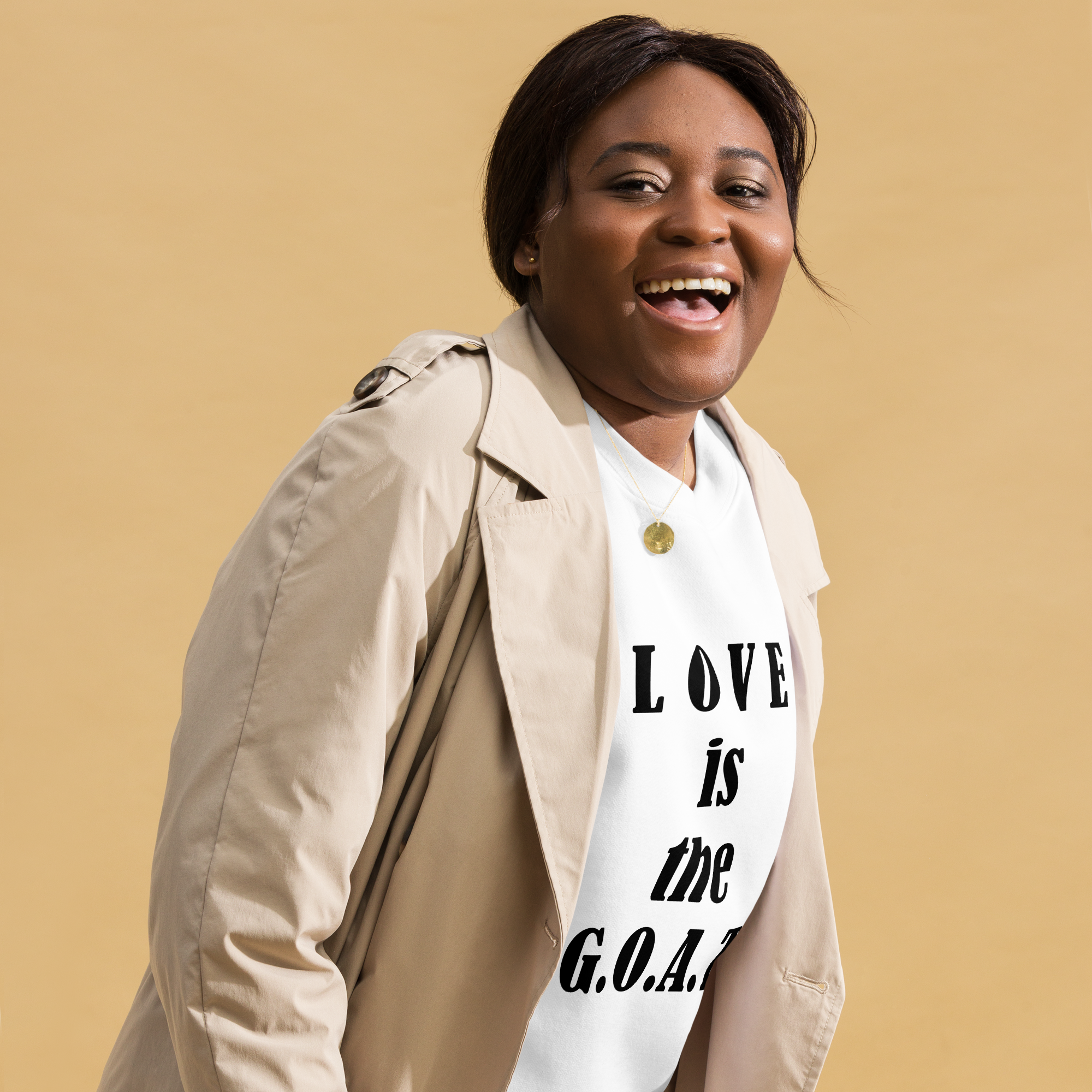 Smiling woman wearing a white shirt with the phrase 'LOVE is the G.O.A.T.' and a beige coat.