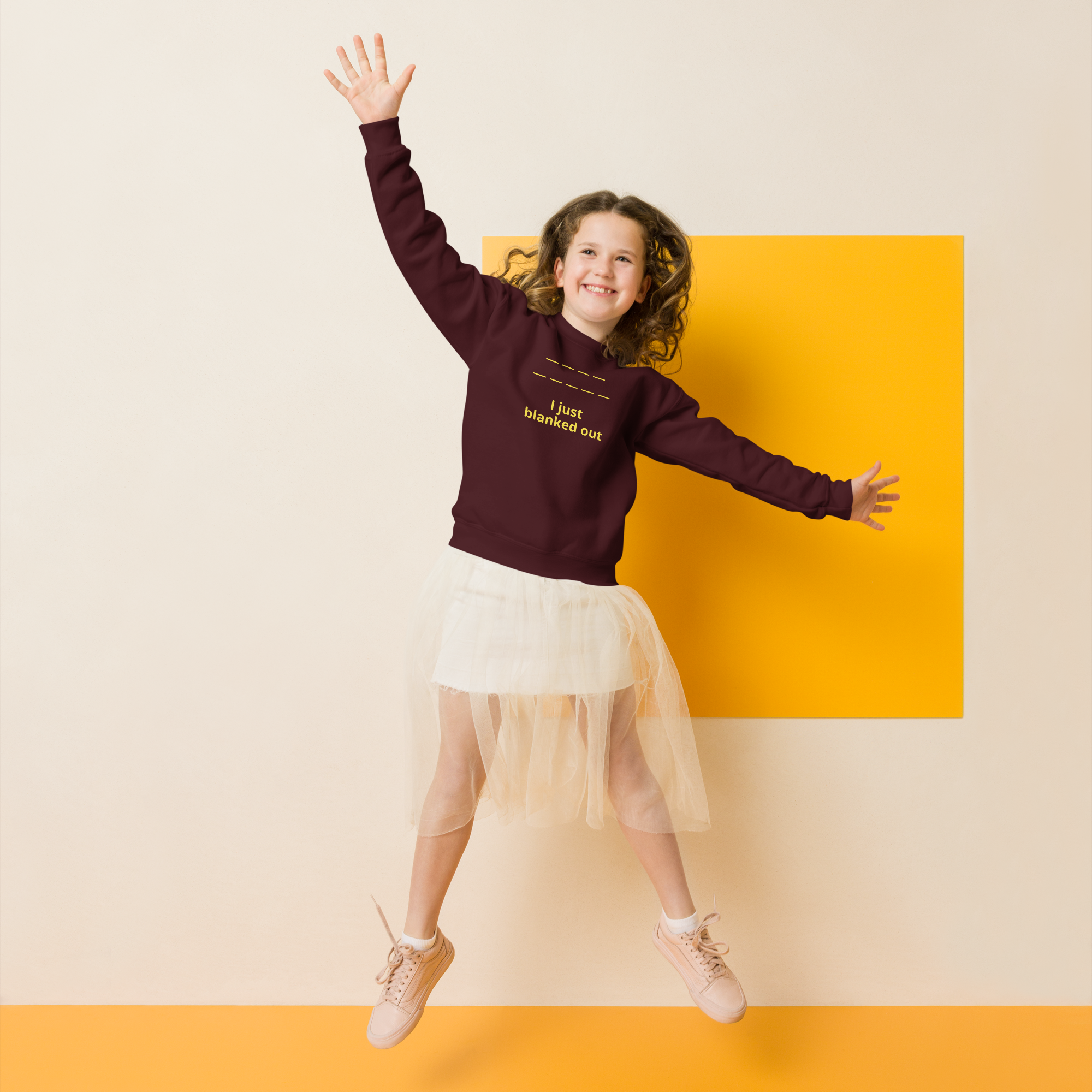 Joyful girl jumping, wearing a maroon sweatshirt and white tulle skirt against a bright yellow backdrop.