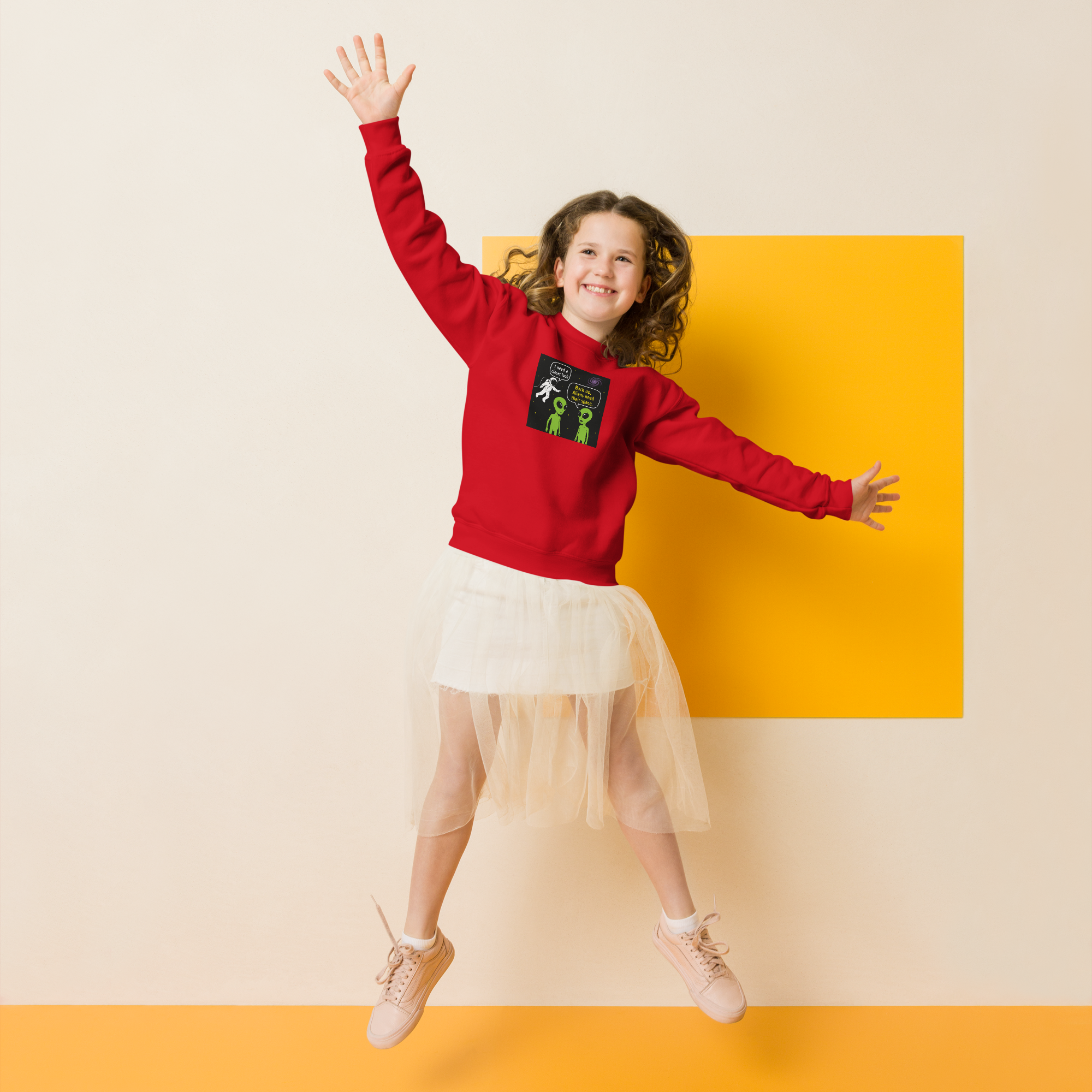 Smiling girl jumping in red sweatshirt with alien cartoon and white skirt.