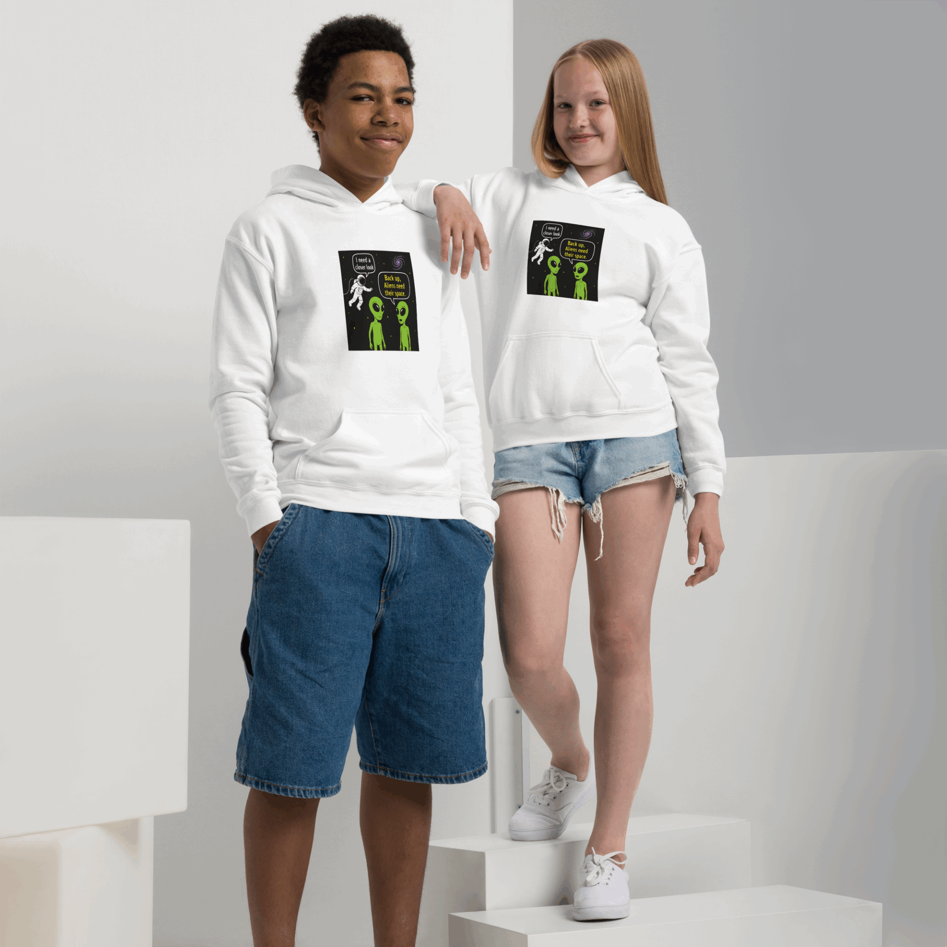 Two kids wear white hoodies with a humorous alien and astronaut comic print.